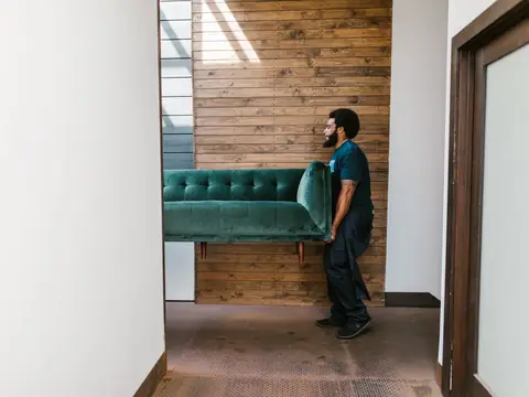 A mover carrying a green sofa through a bright hallway with a wooden wall