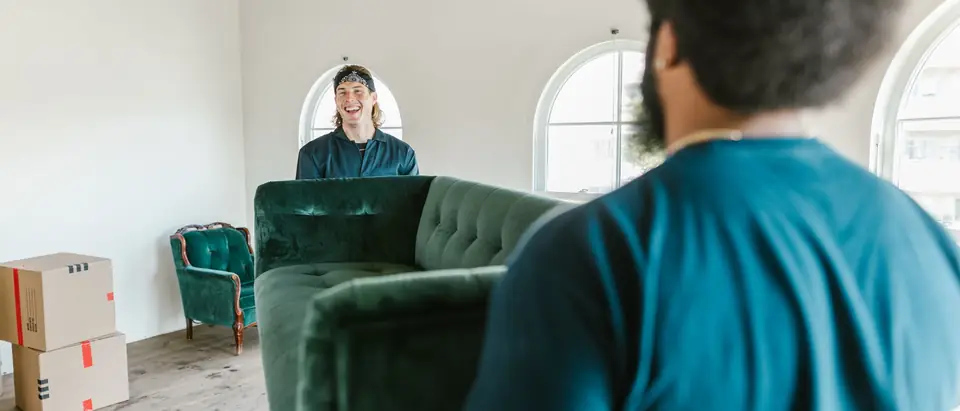 Two movers carrying a green couch through a sunlit room with packed boxes