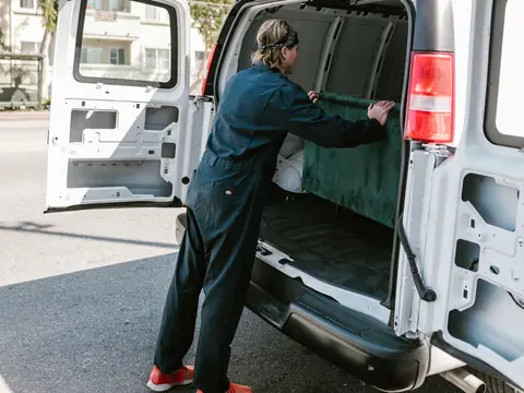 A mover loading a green sofa into the back of a white van on a sunny street