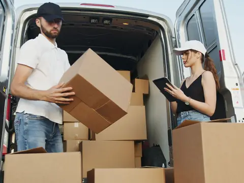 Two workers loading cardboard boxes into the back of a van, one checking a tablet