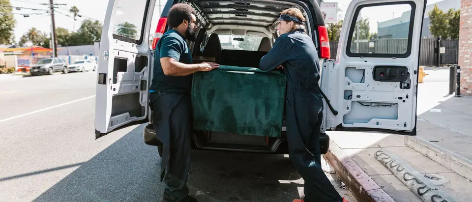 Two movers loading a green sofa into the back of a white van on a city street