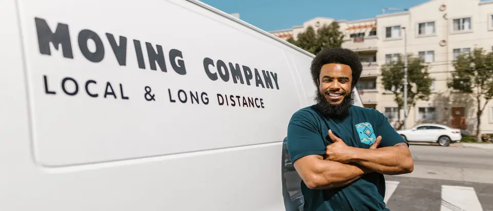 A smiling mover with folded arms beside a white moving van on a sunny street
