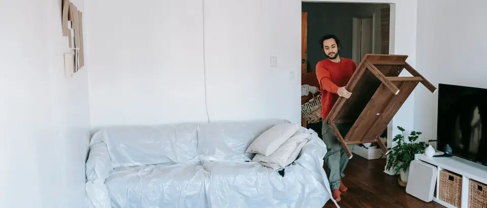 A man carrying a wooden table past a sofa wrapped for moving in a bright room