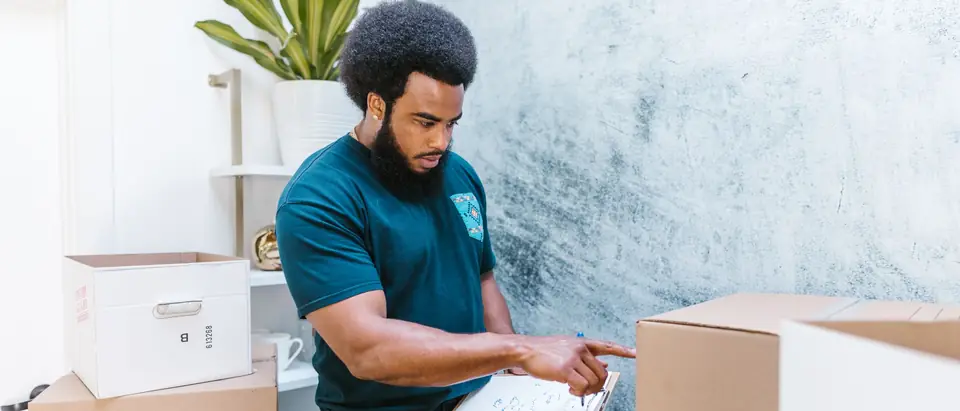 A mover in a teal uniform checking an inventory list beside packed boxes