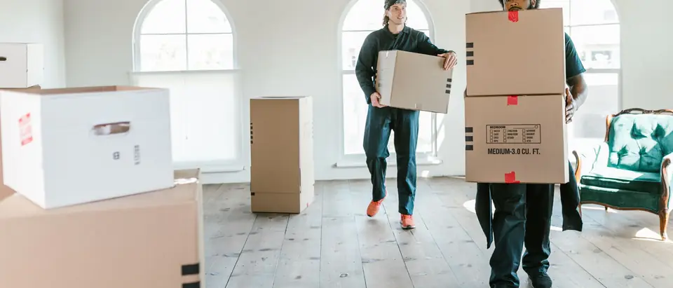 Two movers carrying boxes through a sunlit room with arched windows