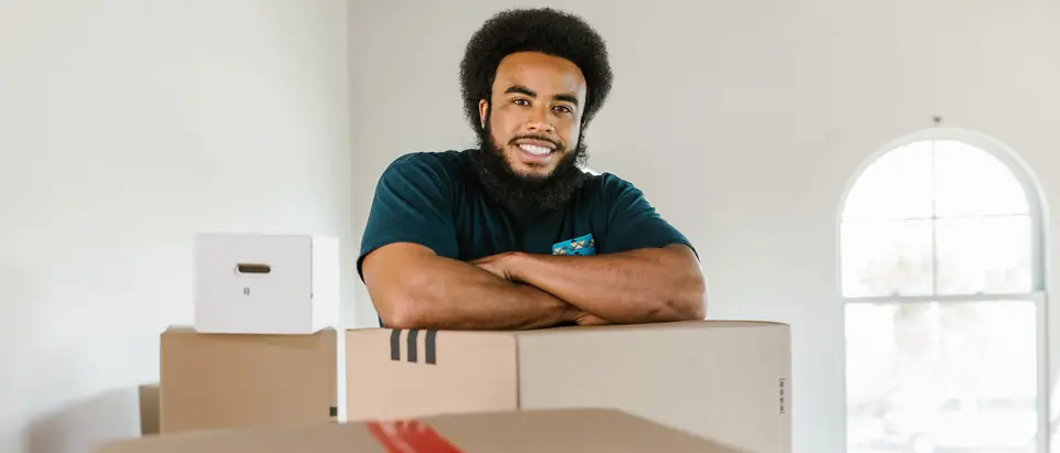A smiling mover leaning on packed moving boxes in a bright room