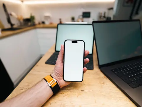 A hand holding a smartphone at a wooden desk beside two laptops