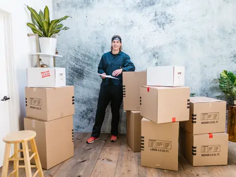 A mover in coveralls holding a clipboard beside stacked moving boxes
