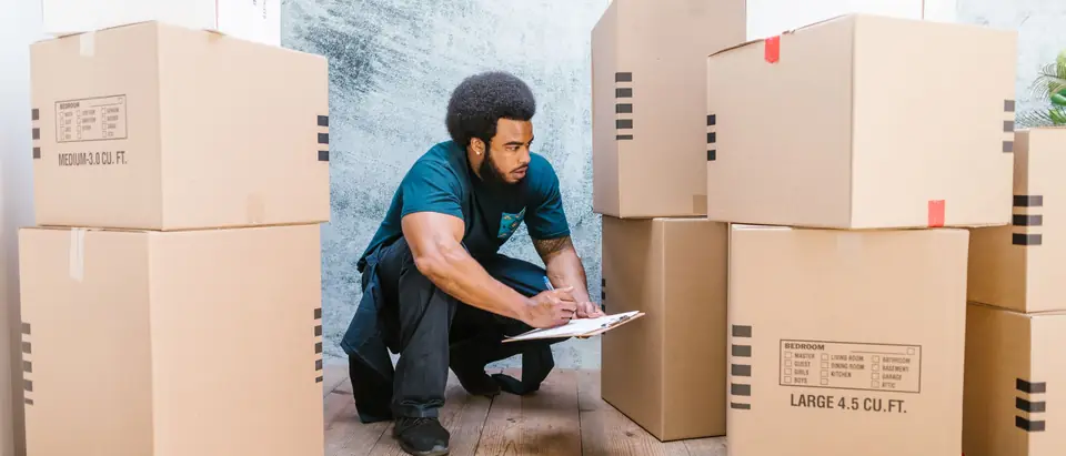 A mover in workwear checking a clipboard among stacked moving boxes